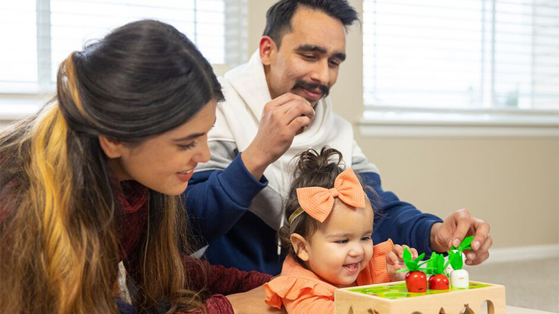 A mother and father happily watch their toddler daughter who is wearing a large orange bow in her hair play with a wooden toy that has carrots inserted in the top