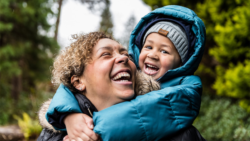 A mother with a huge smile on her face looks up to her left shoulder to see her son, wearing a grey knit hat and a teal hooded puffy jacket, who is wrapping her in a hug from behind.