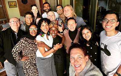 A group of adults participating in the Health Equity Track huddle together looking happy at the camera while standing inside a room with yellow walls and brown doors