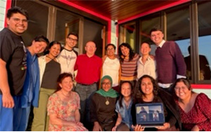 A group of adults participating in the Health Equity Track happily group together, some sitting and some standing, on a porch in from of a white building with red trim and big windows